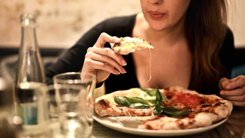 Woman Holds Sliced Pizza Seats by Table With Glass